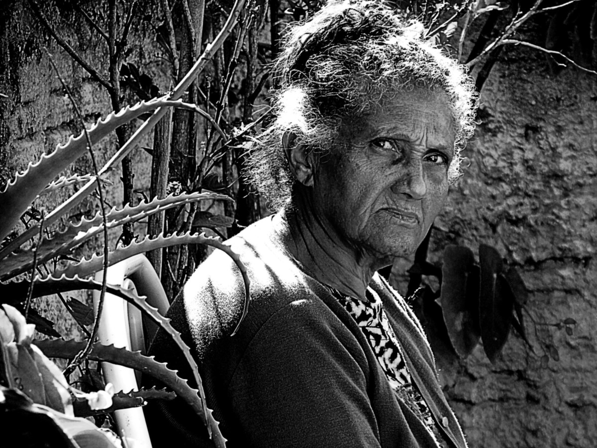 A senhora de cabelos brancos, sentada, observa atentamento a câmera fazer o clique. No seu olhar pensamentos passeiam e, ao redor, o tempo vai passando.
