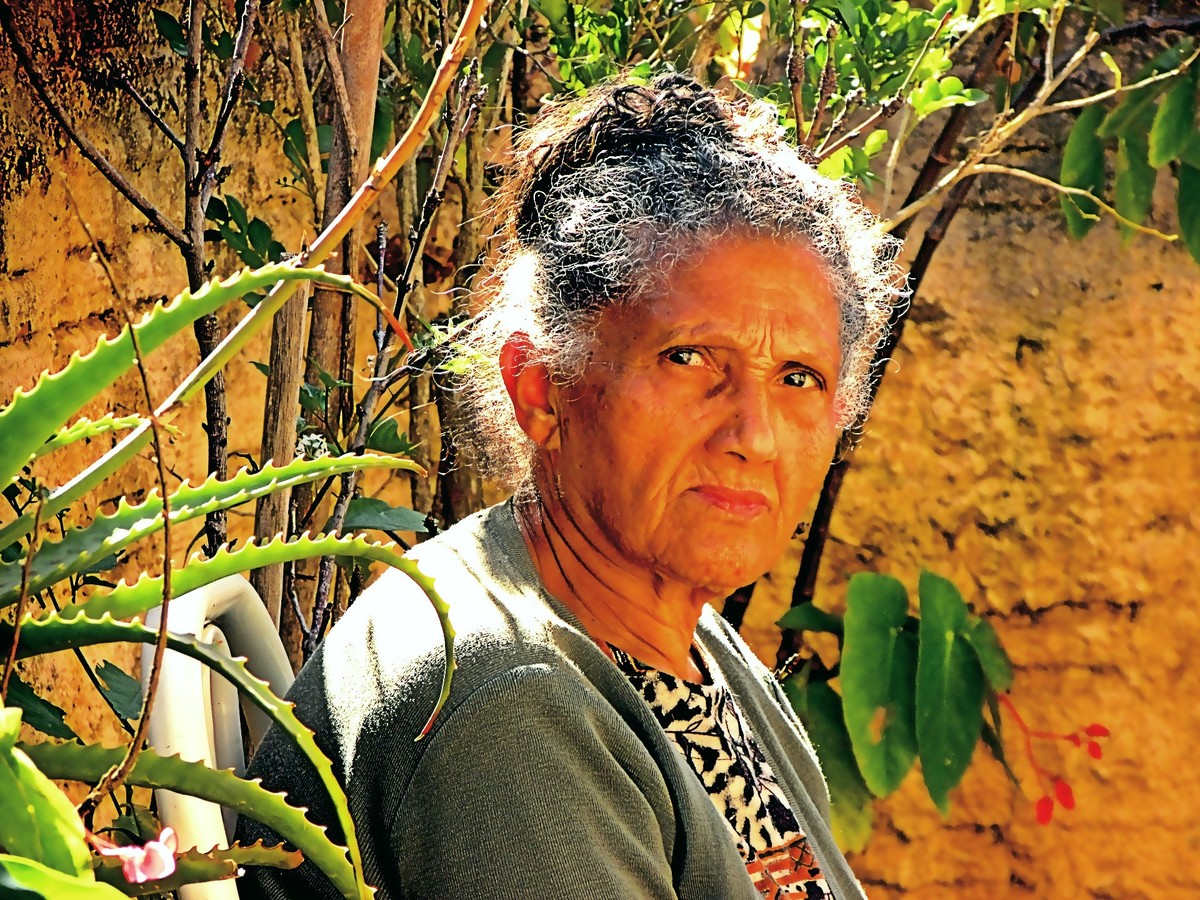 A senhora de cabelos brancos, sentada, observa atentamento a câmera fazer o clique. No seu olhar pensamentos passeiam e, ao redor, o tempo vai passando.