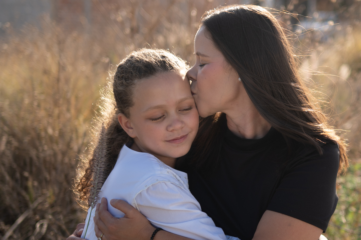 La madre abraza y besa a su hija durante una sesión familiar al atardecer en Querétaro. Una fotografía llena de ternura y conexión emocional.