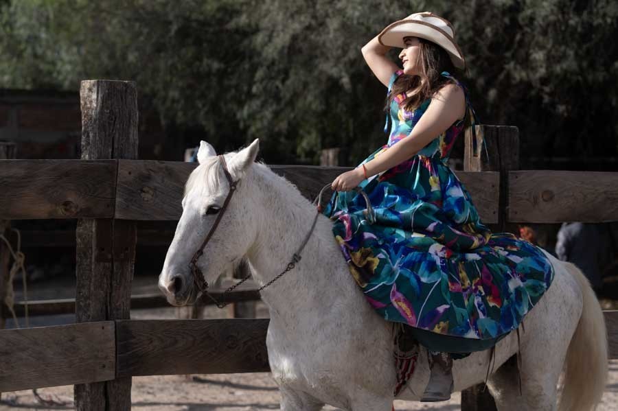 Quinceañera con sombrero vaquero cabalgando un caballo blanco junto a la cerca de madera