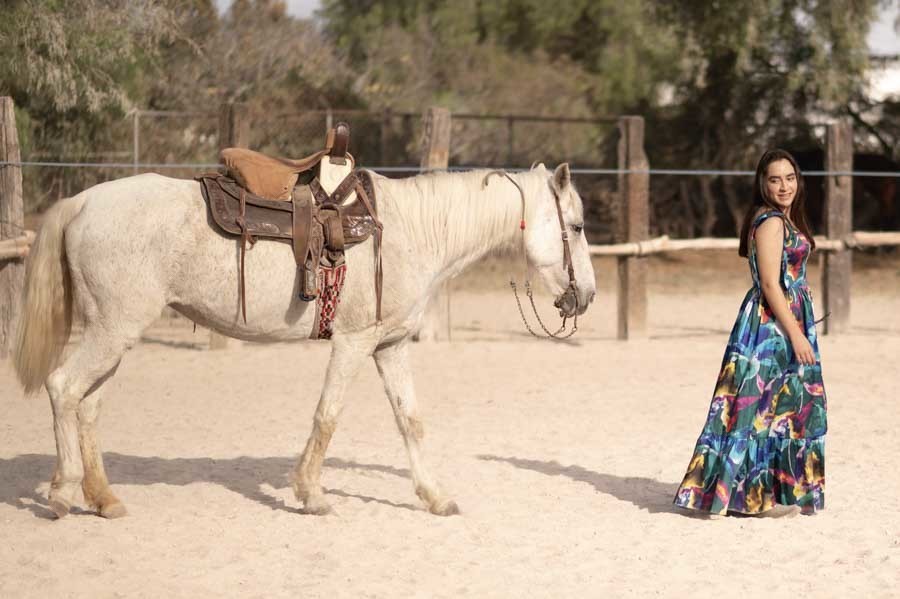 Quinceañera de pie junto a un caballo blanco ensillado en la arena, sesión ecuestre en Querétaro