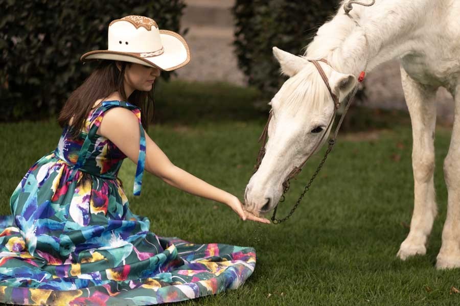 Quinceañera hincada en el pasto dándole de comer a un caballo blanco en su sesión de XV años