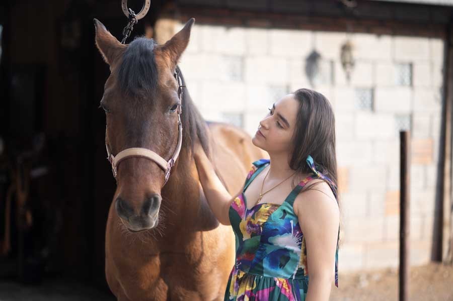 Quinceañera con vestido floral junto a un caballo café en el establo, sesión de XV años
