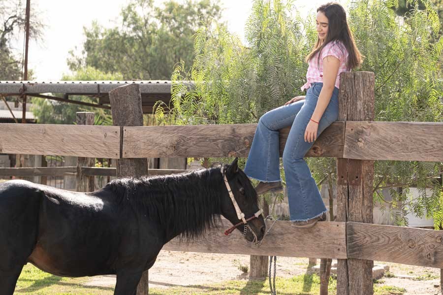 Quinceañera en jeans sentada en una cerca de madera junto a caballos en Querétaro
