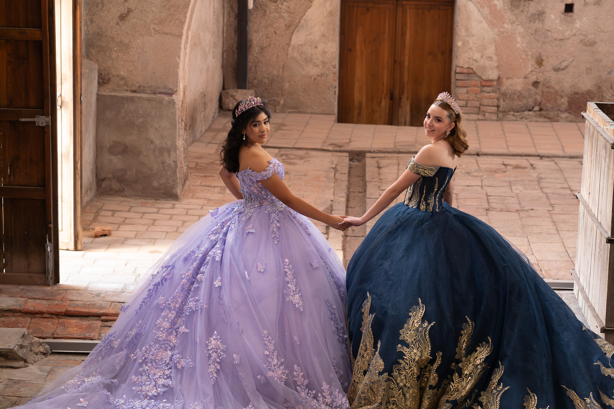 Dos quinceañeras con vestidos lila y azul marino frente a un muro de hacienda