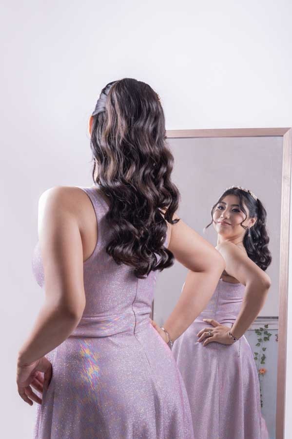 Quinceañera de espaldas frente al espejo, peinado y vestido de XV años lila