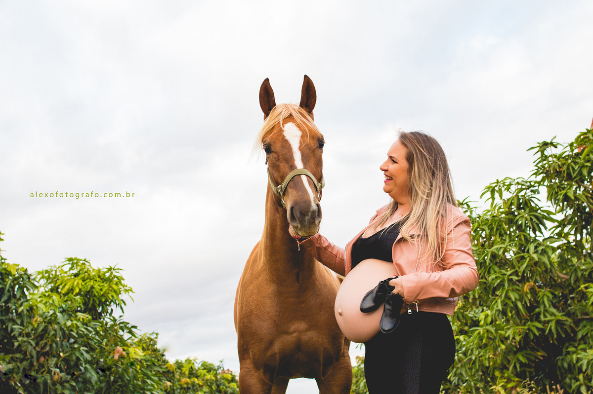 ensaio de gestante no meio da plantação de acerolas vestida de vaqueira segurando uma égua e com a outra mão segurando um par de botinhas