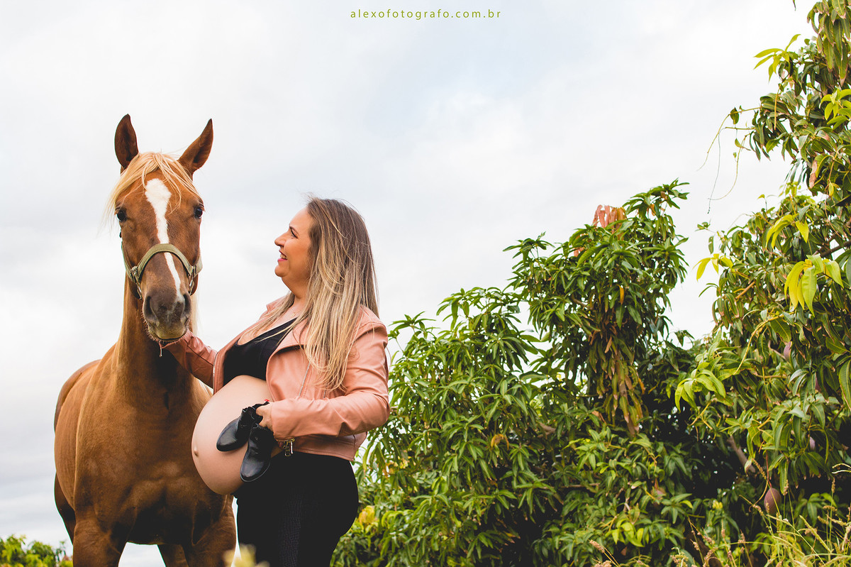 ensaio de gestante no meio da plantação de acerolas vestida de vaqueira segurando uma égua e com a outra mão segurando um par de botinhas sorrindo