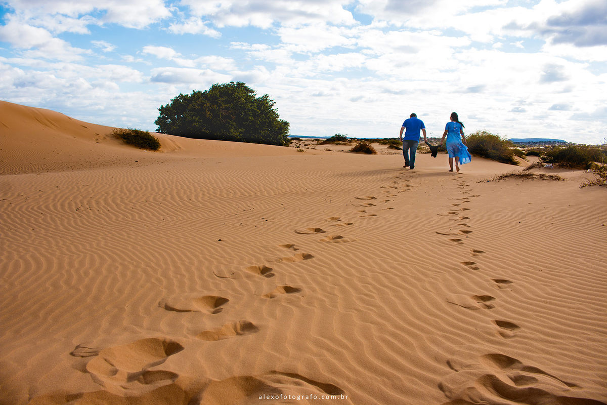 ensaio de gestante, casal caminhando nas areias das dunas de casa nova segurando uma roupinha do bebê pelas mangas, como se estivessem caminhando os três de mãos dadas