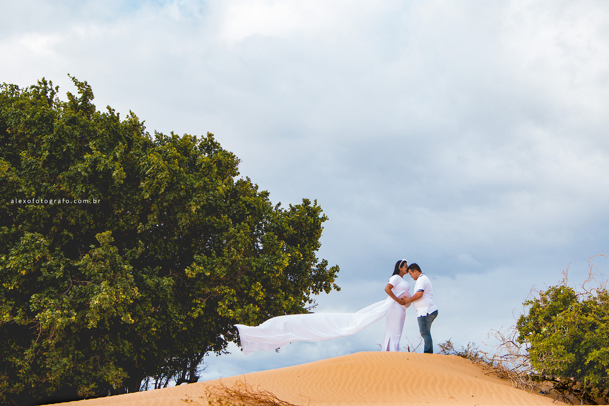 ensaio de gestante, mulher grávida beijando a testa do marido enquanto ele olha para sua barriga enorme e linda, tudo isso numa duna em casa nova ao lado de uma árvore e com o céu nublado ao fundo e o pano do vestido voando