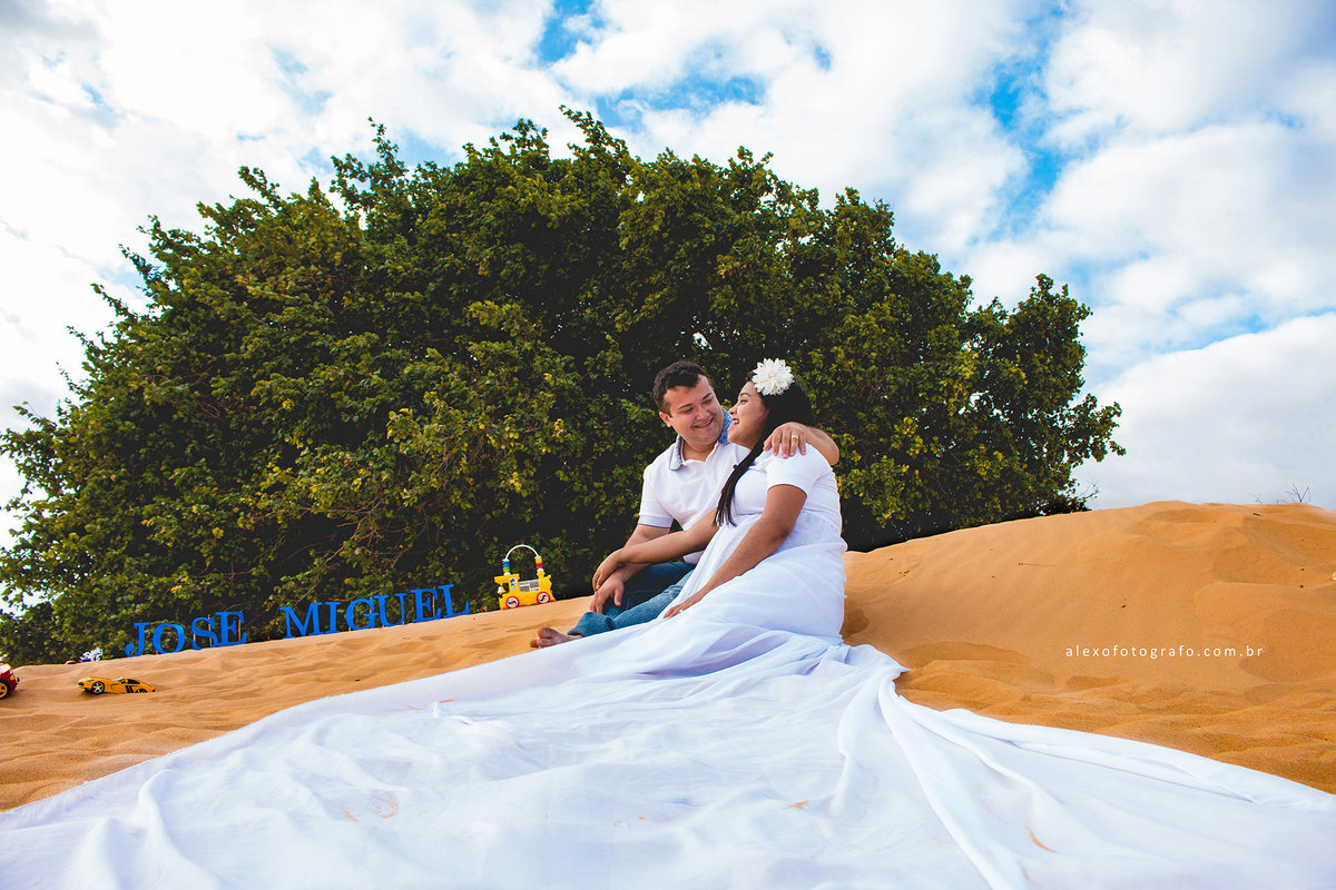ensaio de gestante, casal sentado na areia das dunas de casa nova, o pano do vestido aberto descendo, uma árvore e o céu com muitas nuvens por trás, o nome do José Miguel na areia com alguns brinquedos
