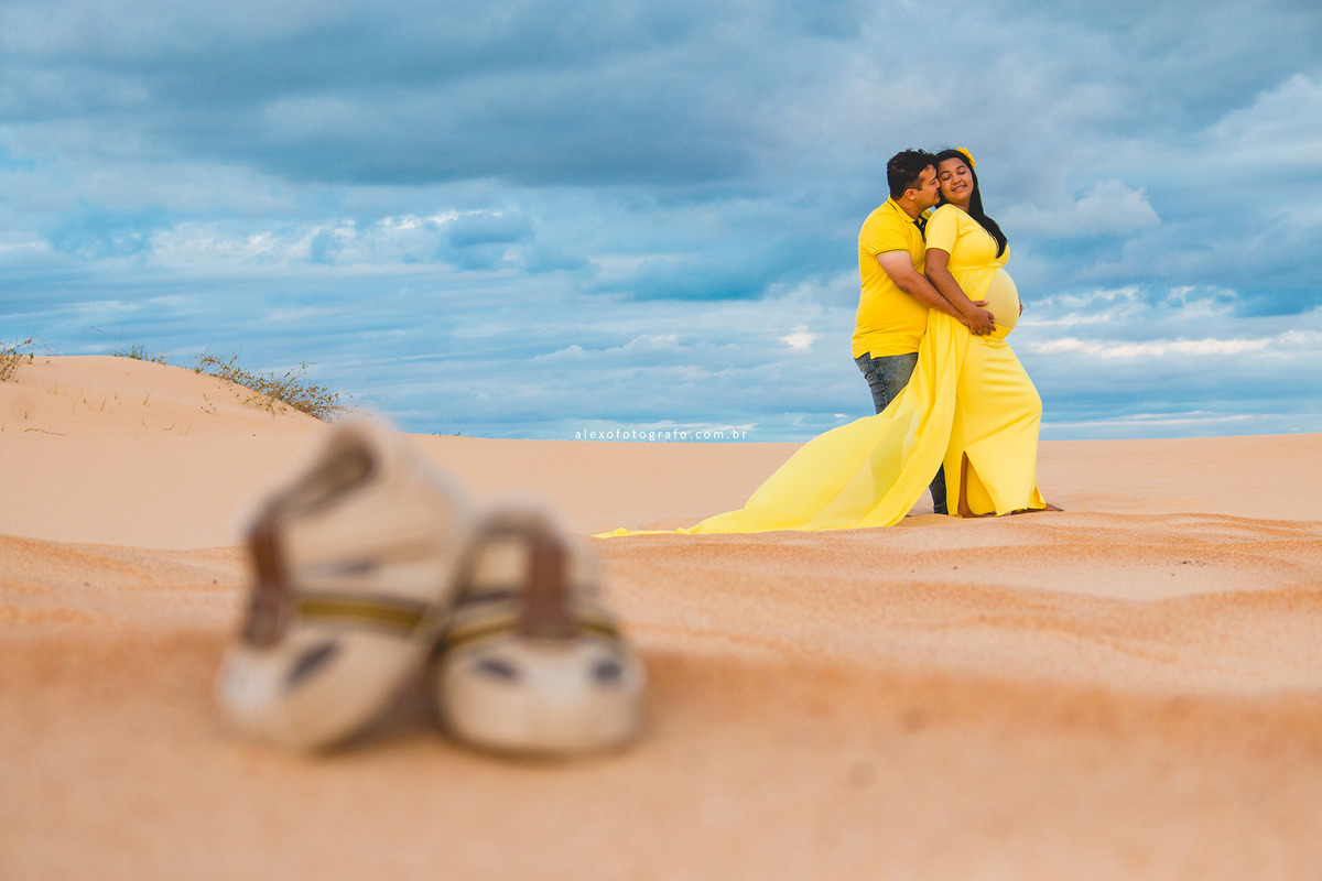 ensaio de gestante, casal abraçado de pé na areia e um par de sapatinhos desfocados na frente próximo
