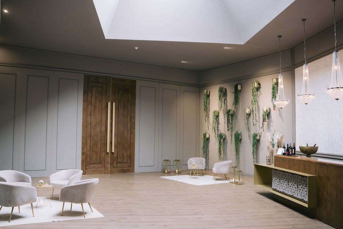 Sala interior com portas de madeira, cadeirões, lustres e parede com plantas
