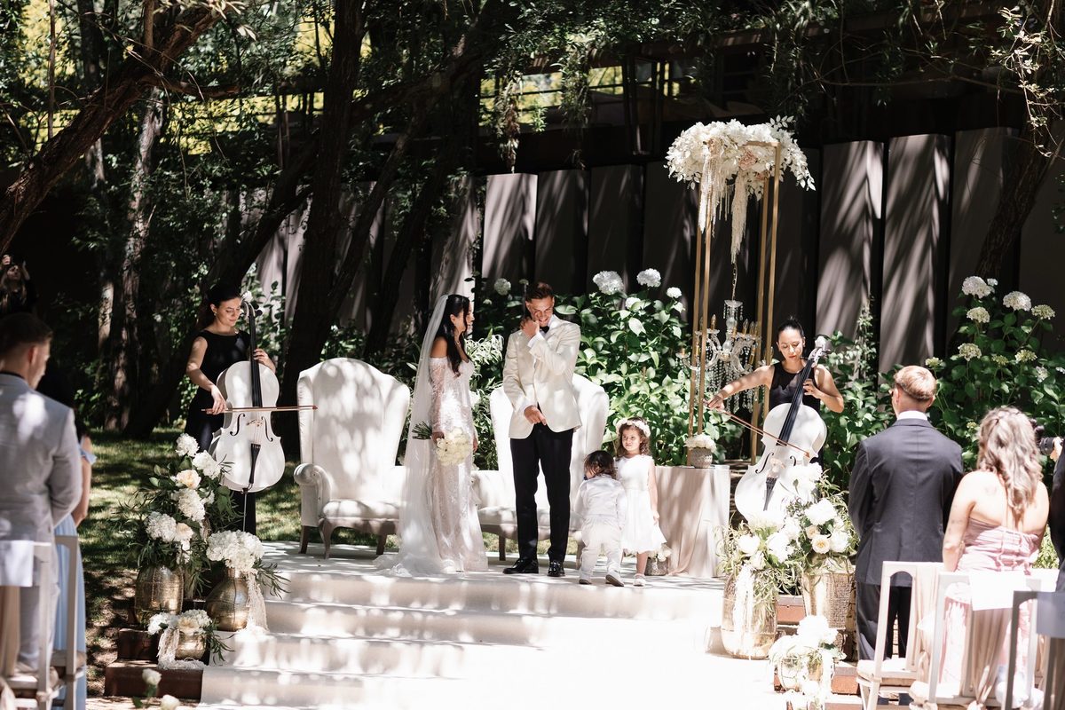 Noivos no altar da cerimónia ao ar livre no bosque, com violoncelistas e arranjos de flores brancas