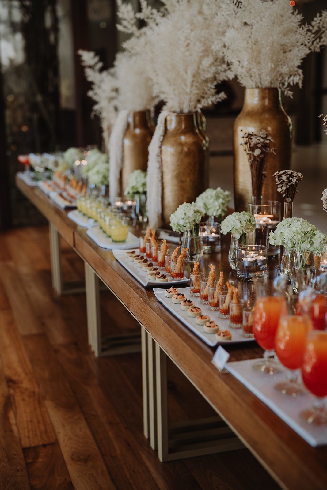 Mesa de cocktail com petiscos, sumos e vasos dourados com flores secas