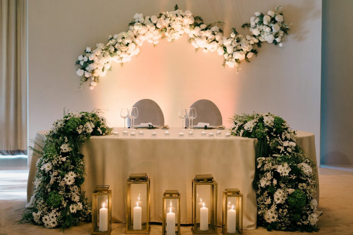 Mesa dos noivos com arco de flores brancas, verdura e lanternas douradas