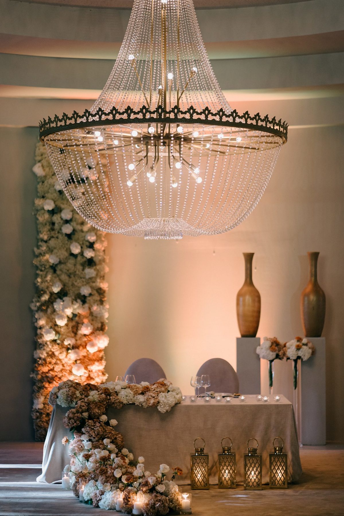 Mesa dos noivos com grande lustre de cristal e parede de flores