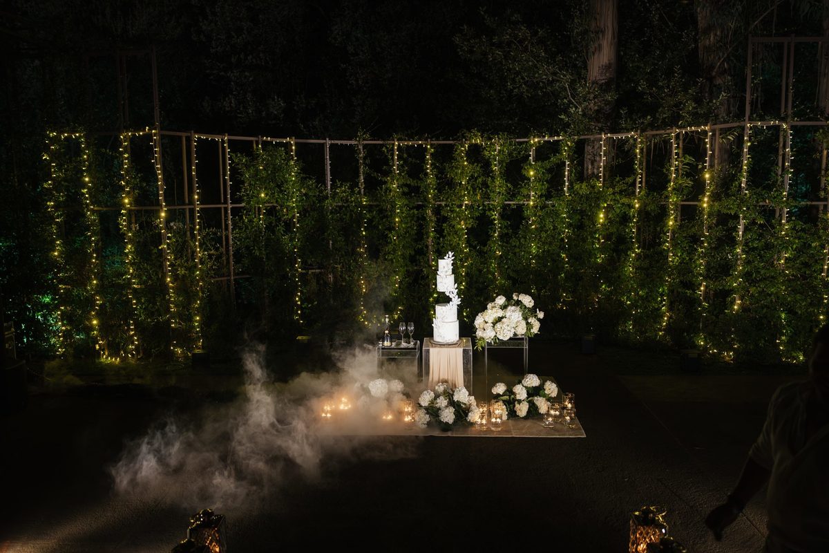 Mesa do bolo no bosque à noite, com cortina de luzes e flores brancas