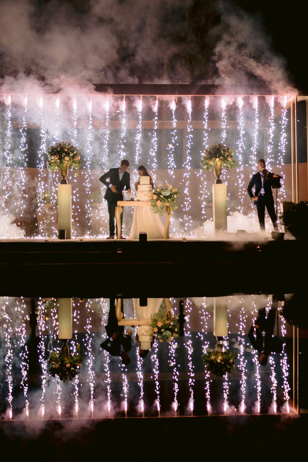 Noivos junto ao bolo com cortina de luzes e reflexo no espelho de água