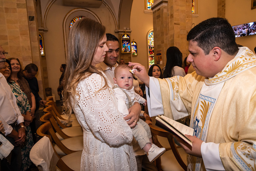 Padre passando óleo na testa do Antonio que está no colo da mãe e olhando para a Câmera - FOTOGRAFIA DE BATIZADO - PARÓQUIA NOSSA SENHORA APARECIDA DE MOEMA - FOTÓGRAFO DE BATIZADO EM SÃO PAULO - RODRIGO MARTIN FOTOGRAFIA