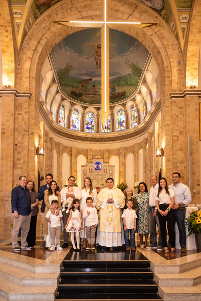 Toda a família do Antonio reunida com o padre no altar, mostrando a grande cruz iluminada - FOTOGRAFIA DE BATIZADO - PARÓQUIA NOSSA SENHORA APARECIDA DE MOEMA - FOTÓGRAFO DE BATIZADO EM SÃO PAULO - RODRIGO MARTIN FOTOGRAFIA