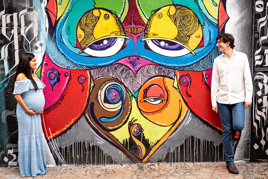 Victoria de lado para a foto e Samuel de frente, ambos se olhando e no meio deles um grafite com alguns olhos - ENSAIO GESTANTE - BECO DO BATMAN - PARQUE IBIRAPUERA - FOTÓGRAFO PROFISSIONAL - FOTÓGRAFO DE GESTANTE - GRÁVIDAS - RODRIGO MARTIN FOTOGRAFIA