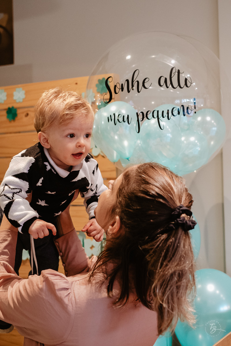 mãe erguendo seu filho no aniversário de primeiro ano, com balão ao fundo com frase escrita: Voe alto meu pequeno.