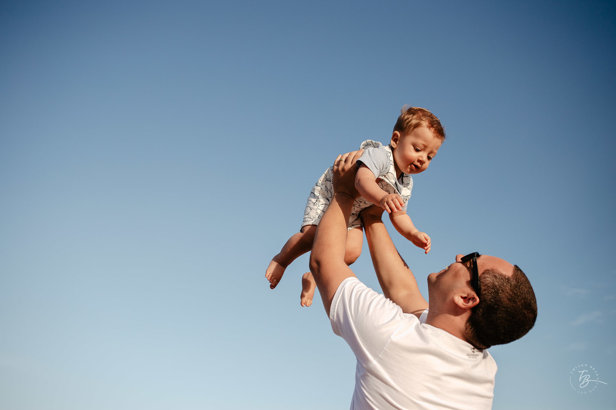 Bebê erguido por seu pai na praia em dia de céu azul Ensaio lifestyle. Fotos de Família em Garopaba - SC. Por Thiago Braga
