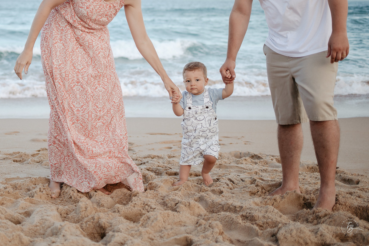 Criança pequena de mãos dadas com seus pais, na praia do Silveira. Ensaio lifestyle. Fotos de Família em Garopaba - SC. Por Thiago Braga