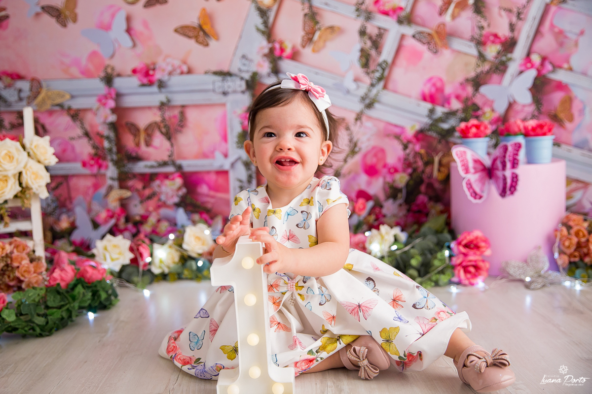 Foto de smash the cake no cenário jardim e borboletas, menina com vestido florido segurando o número um.