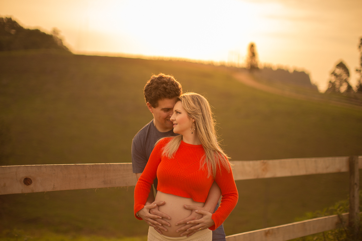 Ensaio gestante da Pamela e Fabio com Gustavo Sousa fotografia em Curitiba. Book em externa em haras. Gravida com calça amarela e blusa laranjada. Pose na cerca com por-do-sol 
