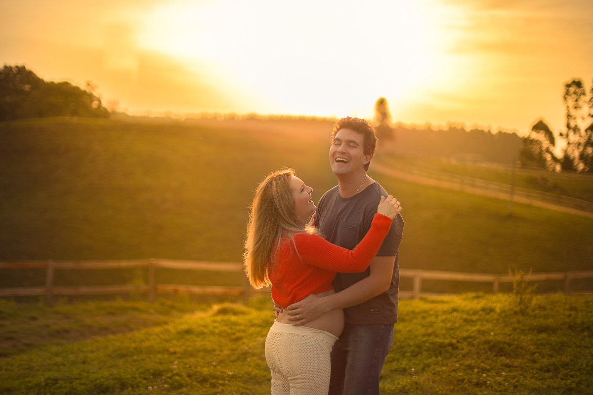 Ensaio gestante da Pamela e Fabio com Gustavo Sousa fotografia em Curitiba. Book em externa em haras. Gravida com calça amarela e blusa laranjada. Pose na cerca com por-do-sol em foto expontânea com risada