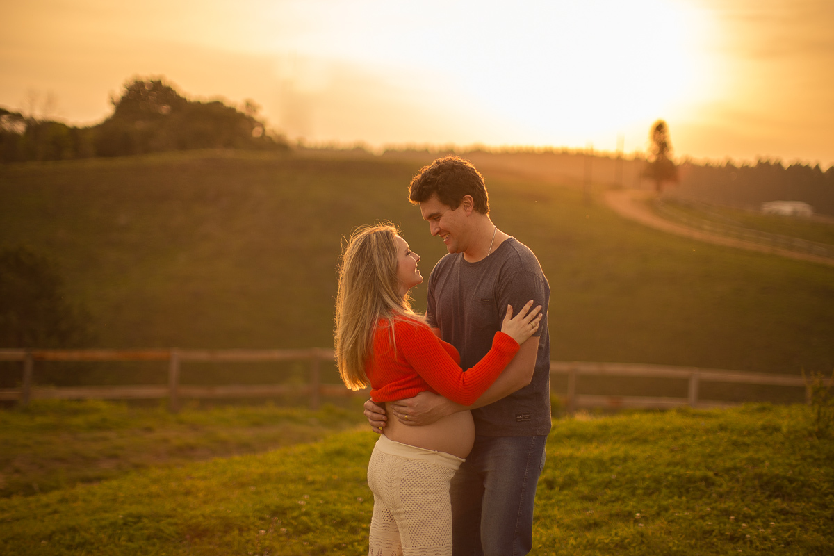 Ensaio gestante da Pamela e Fabio com Gustavo Sousa fotografia em Curitiba. Book em externa em haras. Gravida com calça amarela e blusa laranjada. Pose na cerca com por-do-sol em foto expontânea apaixonados