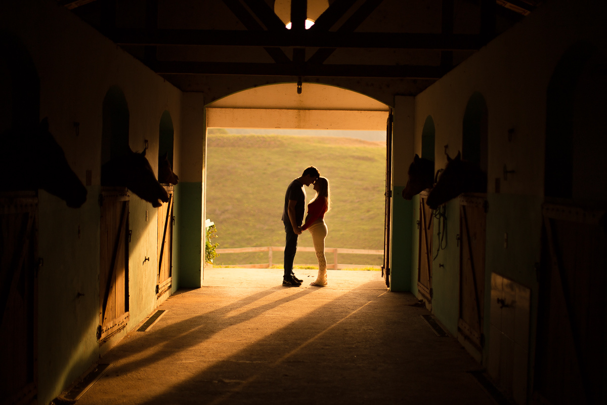 Ensaio gestante da Pamela e Fabio com Gustavo Sousa fotografia em Curitiba. Book em externa em haras. Gravida com calça amarela e blusa laranjada. Pose de frente pra o outro no coxo dos cavalos