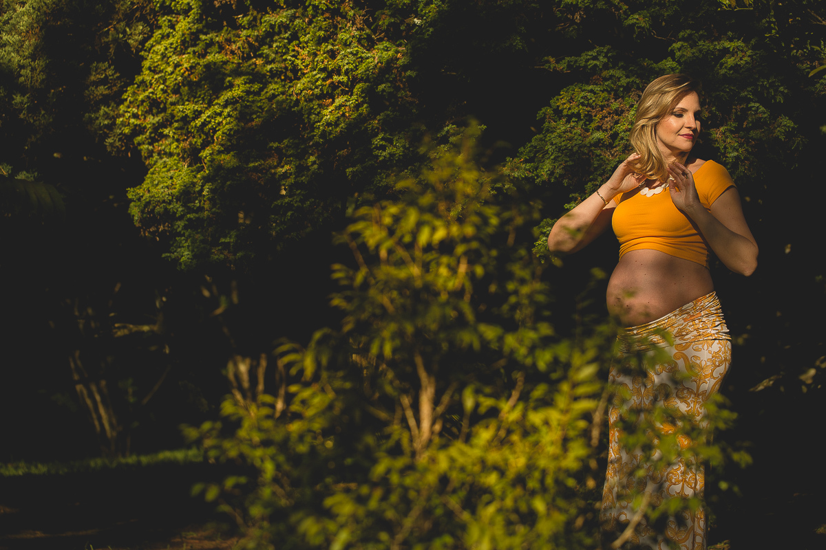 Ensaio gestante da Mariana e Zé com Gustavo Sousa fotografia em Curitiba. Book em externa. Blusa Laranja com calça florida laranja e branco. Foto em meio ao jardim. Sol
