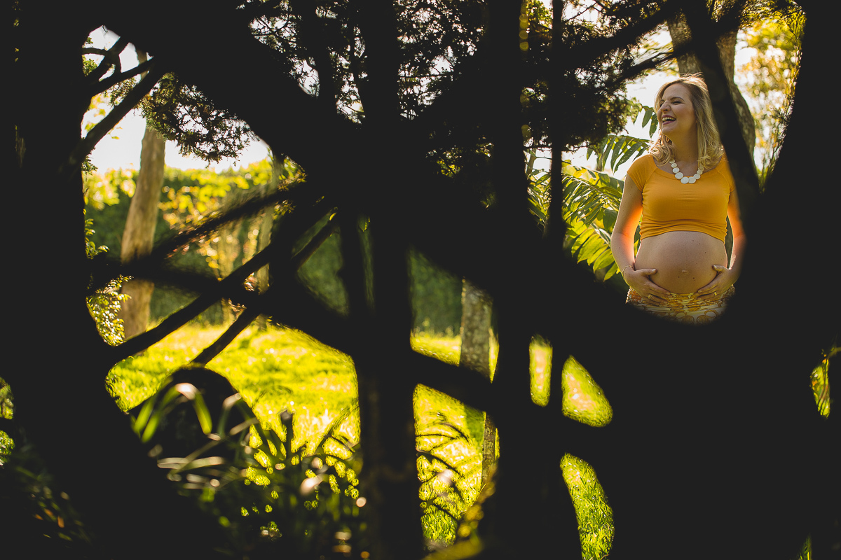Ensaio gestante da Mariana e Zé com Gustavo Sousa fotografia em Curitiba. Book em externa. Blusa Laranja com calça florida laranja e branco. Foto em meio ao jardim com mão na barriga