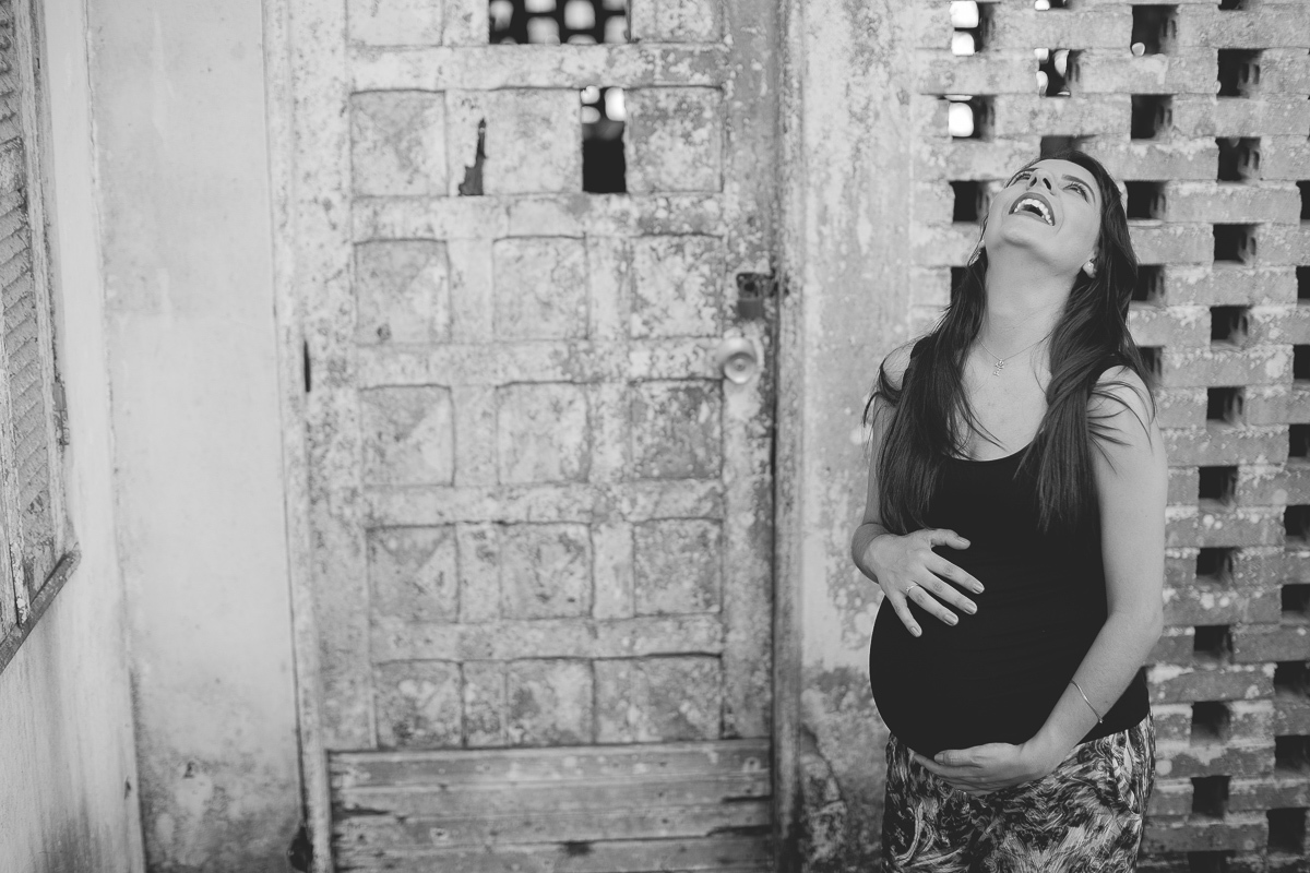ensaio gestante em externa feito em curitiba com Gustavo Sousa Fotografia. Gesntante dando rizada em frente ao muro vazado com porta velha. Foto em preto e branco.