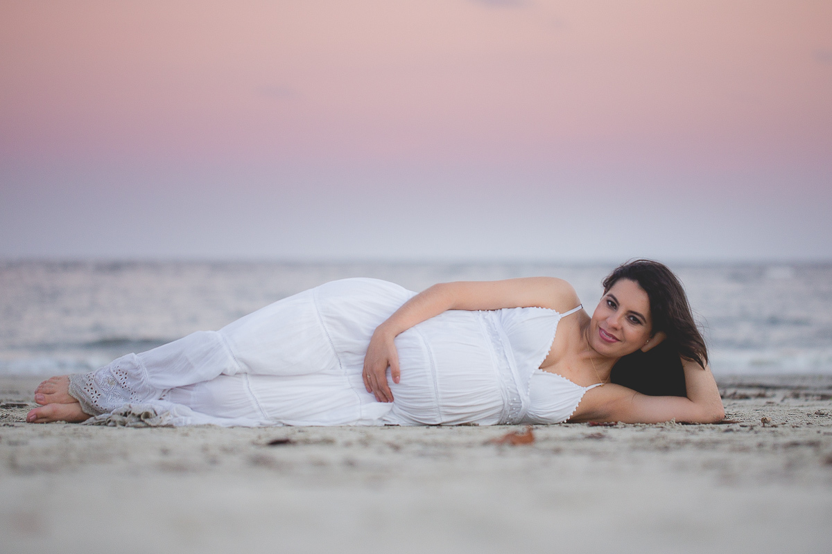 ensaio gestante em externa feito em curitiba com Gustavo Sousa Fotografia. Foto na praia com gestante no colo. por do sol. gestante de vestido branco deitada na areia.