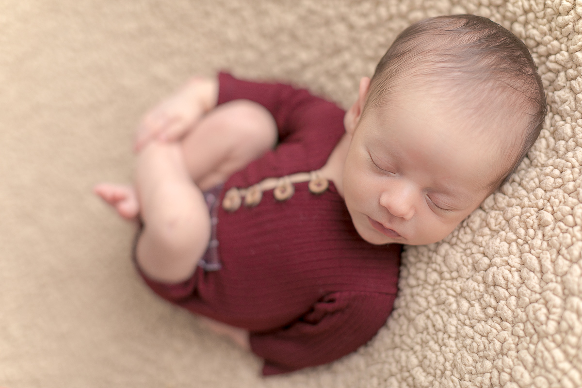 Foto feita no estúdio Gustavo Sousa Newborn em Curitiba. Bocejo do bebê com roupinha vermelha com botões de madeira. Fundo de manta bege. Pose de barriguinha para cima.