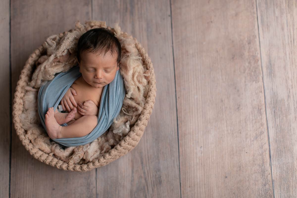 Ensaio Newborn do Theo feito no estúdio Gustavo Sousa fotografia em Curitiba. Fundo de madeira. Enrolado no wrap azul. Pelo e, volta do acessório de crochet. Pose barriga para cima. Fundo de madeira. Foto de cima.