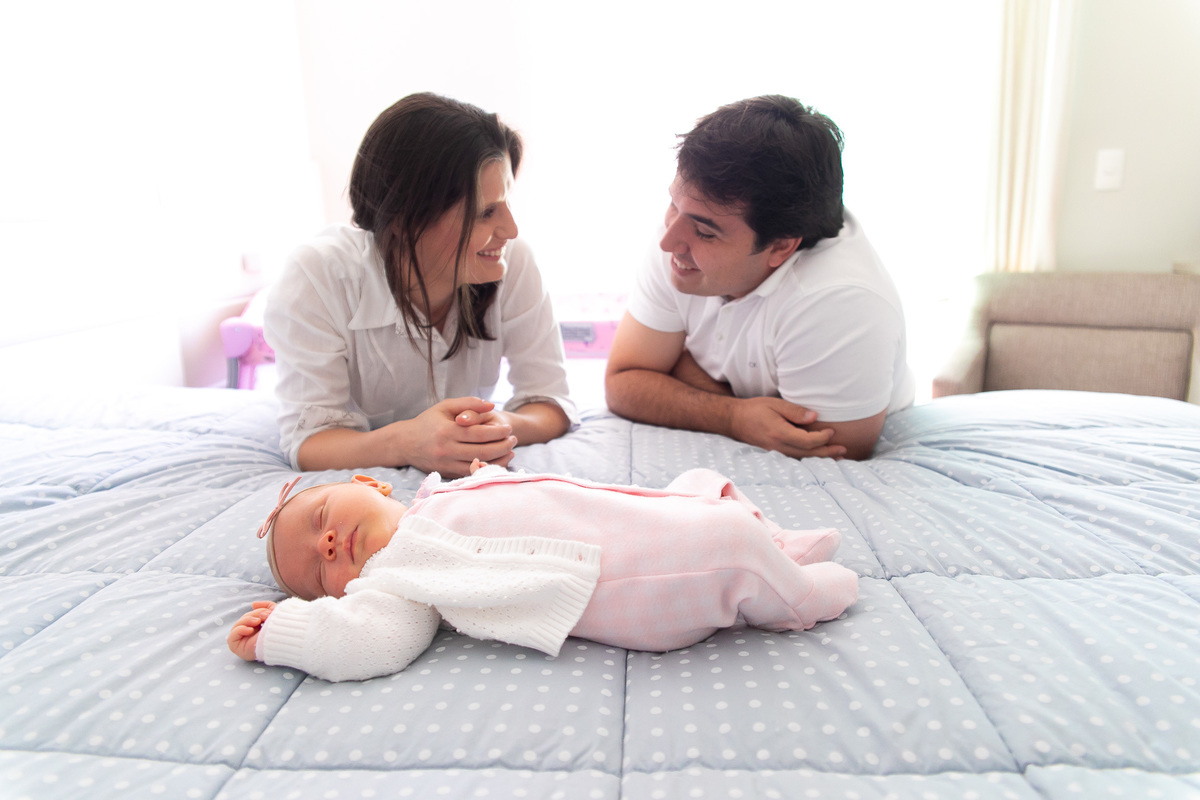 Gustavo Sousa estúdio de fotografia em Maringá-PR. Ensaio e book fotográfico newborn lifestyle e família.. Foto em família, pais se olhando e sorrindo e bebe dormindo na cama.