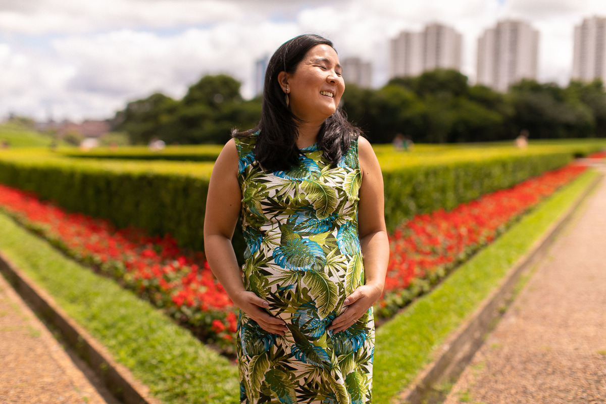 Gustavo Sousa estúdio de fotografia em Maringá-PR. Ensaio e book fotográfico gestante. Foto de grávida com as mãos na barriga sorrindo em um jardim.