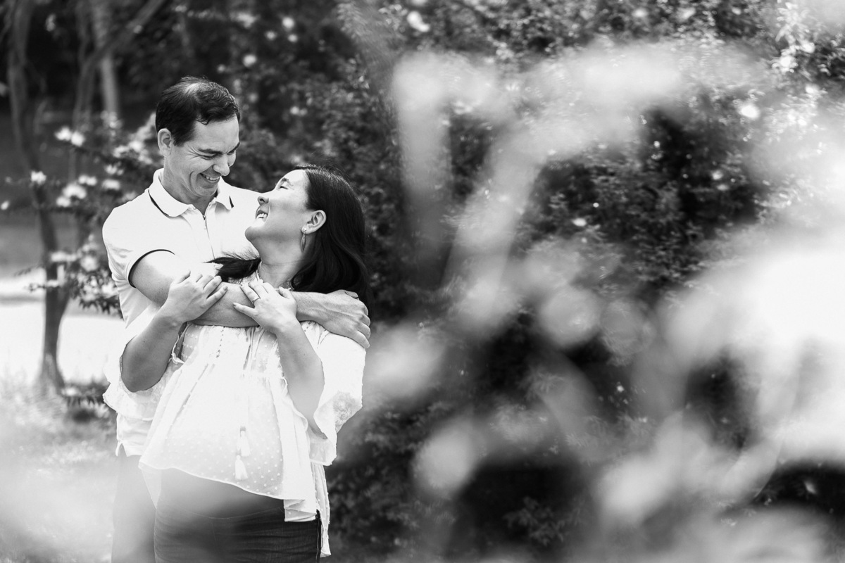 Gustavo Sousa estúdio de fotografia em Maringá-PR. Ensaio e book fotográfico gestante. Foto pai abraçando mãe grávida, casal sorrindo e se olhando, preto e branco.
