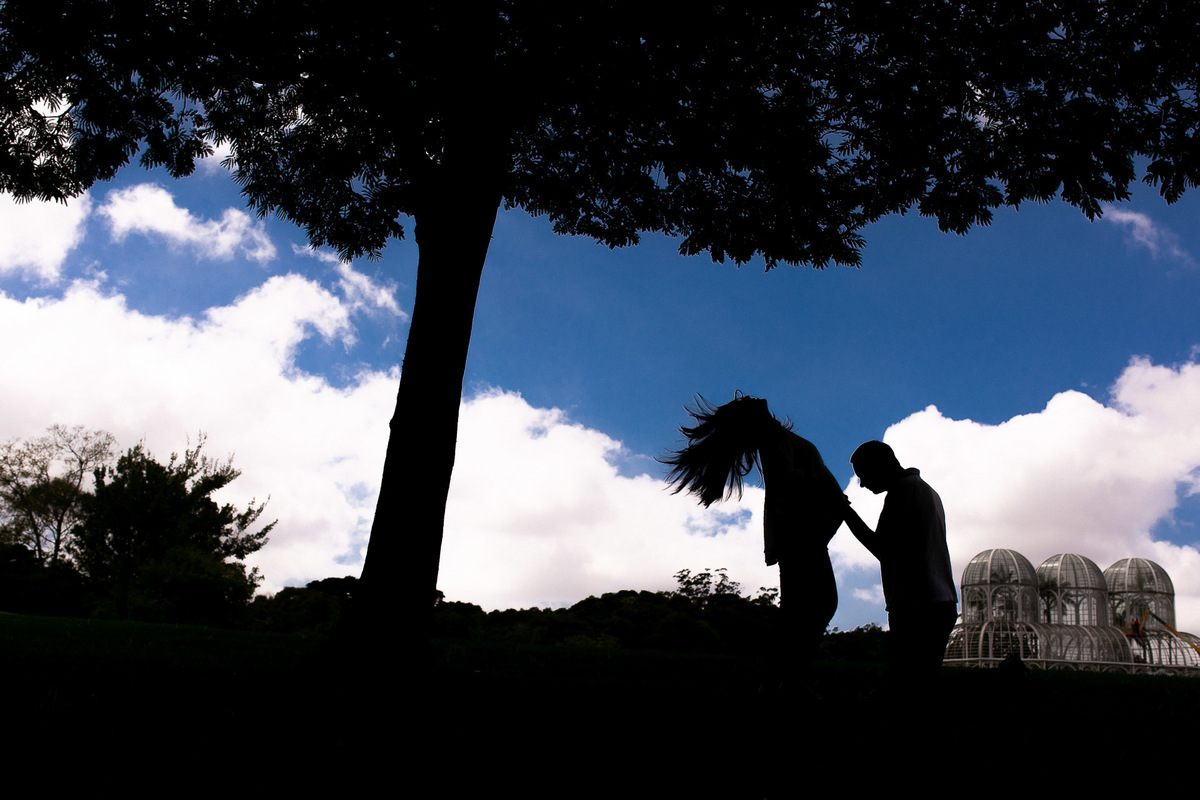Gustavo Sousa estúdio de fotografia em Maringá-PR. Ensaio e book fotográfico gestante. Foto de silhueta, gestante e pai pegando na barriga com estufa do Jardim Botânico de Curitiba ao fundo.