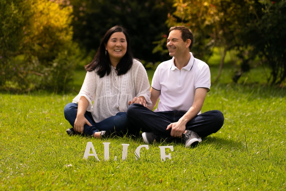 Gustavo Sousa estúdio de fotografia em Maringá-PR. Ensaio e book fotográfico gestante. Foto casal sentados na grama sorrindo, gestante, com o nome Alice na frente.
