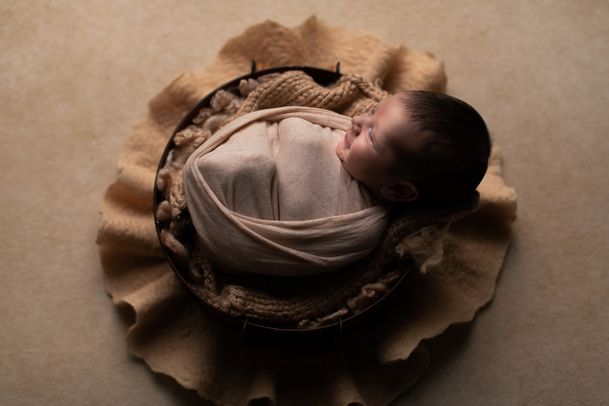 Gustavo Sousa estúdio de fotografia em Maringá-PR. Ensaio e book fotográfico newborn.  Foto bebê enrolado em tecido wrap bege em cesto com fundo neutro.