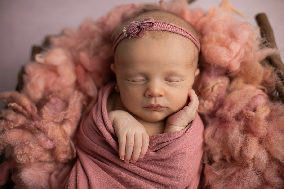 Gustavo Sousa estúdio de fotografia em Maringá-PR. Ensaio e book fotográfico newborn.  Foto bebê recém nascida em cesto enrolada em tecido wrap rosa com a mão no rosto