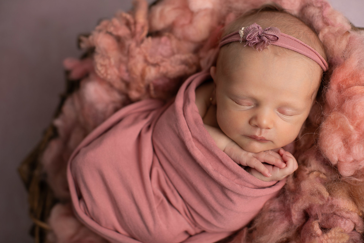 fotográfico newborn.  Foto bebê recém nascida em cesto enrolada em tecido wrap rosa com a mão no queixo