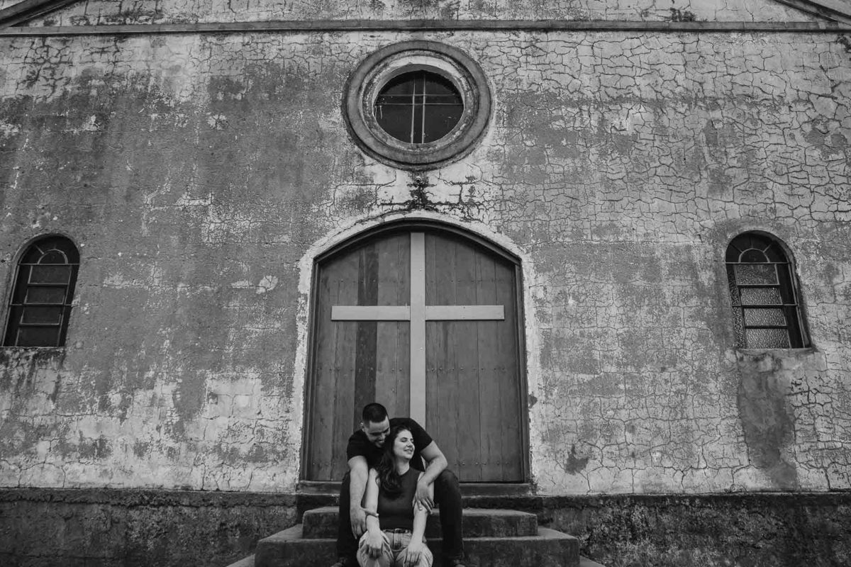 Capela Sagrada Família - Ruínas - em Mandaguari, região de Maringá. Casal vestidos de preto em frente a porta da igreja. Foto por Gustavo Sousa Fotografia. Casal na frente da igreja