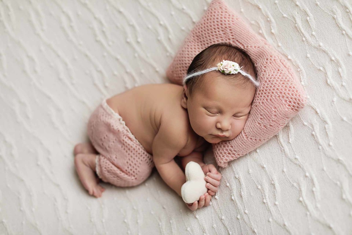 Gustavo Sousa fotografia ensaio de recém-nascido newborn em Maringá. Bebê deitado em manta branca com travesseiro rosa. Roupinha rosa. Coração de pelúcia.
